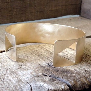 May include: A gold-toned metal cuff bracelet with a geometric design. The bracelet has two cut-out triangles on either side.