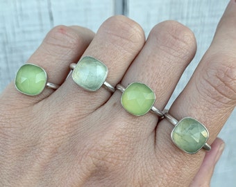 May include: Four silver rings with square green gemstones. The rings are stacked on a person's fingers.