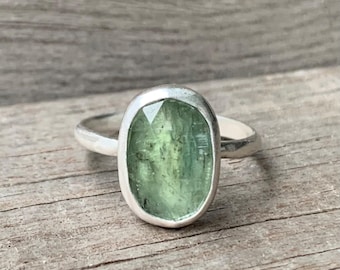 May include: A silver ring featuring a faceted, oval-shaped light green gemstone set in a smooth bezel. The stone displays natural inclusions and a translucent quality, while the band is a simple, polished silver wire that encircles the finger.