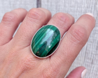 May include: A large oval ring featuring a polished green malachite stone with dark green stripes. The stone is set in a silver-colored band. The ring is shown on a hand.