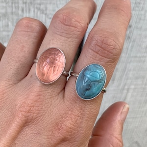 May include: Two silver rings with oval-shaped stones. The stone on the left ring is a light pink color and the stone on the right ring is a light blue color. Both stones have a carved scarab beetle design.