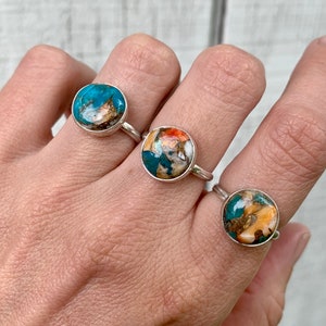 May include: Three silver rings with round turquoise, orange, and brown gemstone settings. The rings are on a person's hand.
