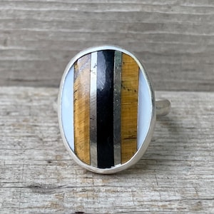 May include: Oval-shaped silver ring with a mosaic design. The mosaic features vertical stripes of white, black, tiger's eye, and silver. The ring is set against a weathered wooden surface.
