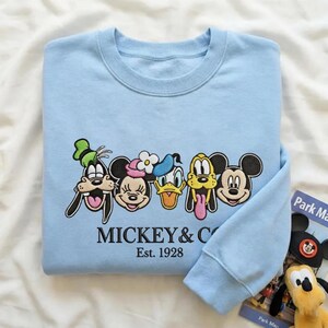 以下が含まれることがあります： グーフィー、ミニーマウス、ドナルドダック、プルート、ミッキーマウスなど、刺繍された漫画のキャラクターの顔が特徴のライトブルーのスウェットシャツ。「MICKEY & CO」と「Est. 1928」の文字が刺繍されています。