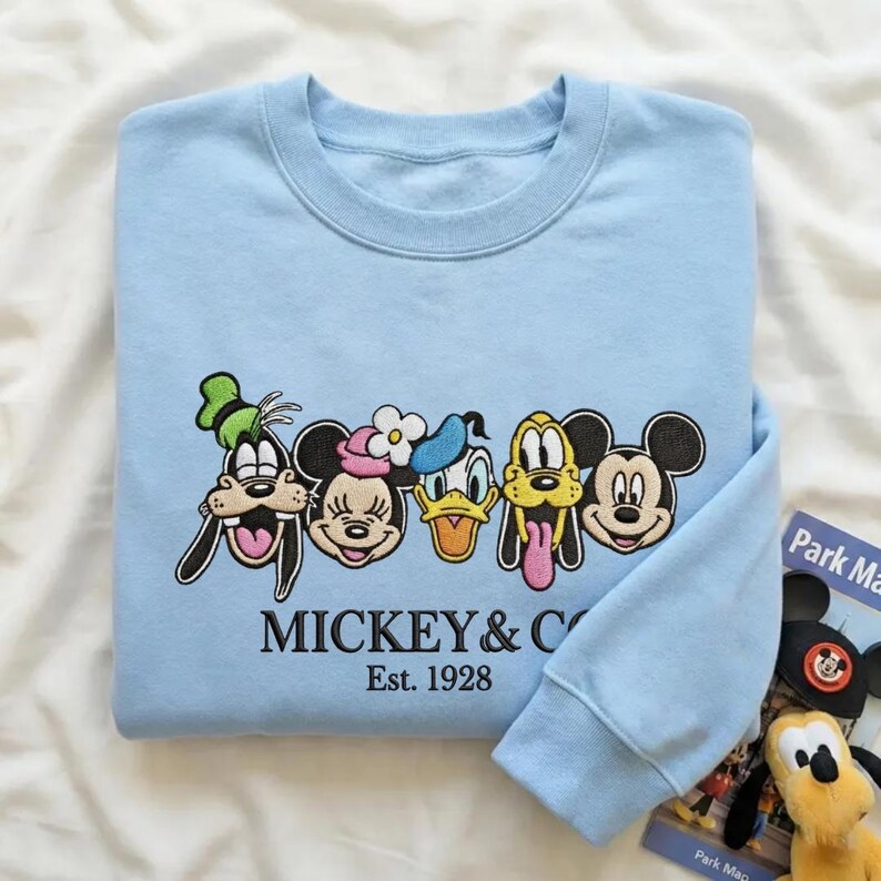 以下が含まれることがあります： グーフィー、ミニーマウス、ドナルドダック、プルート、ミッキーマウスなど、刺繍された漫画のキャラクターの顔が特徴のライトブルーのスウェットシャツ。「MICKEY & CO」と「Est. 1928」の文字が刺繍されています。