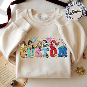 May include: Cream-colored sweatshirt featuring the word "CUSTOM" in colorful letters, each letter adorned with a Disney princess. The design includes Belle, Jasmine, Rapunzel, Snow White, Ariel, and Elsa. The sweatshirt has long sleeves.