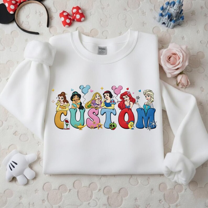 May include: White sweatshirt with a colorful graphic featuring Disney princesses and the word "CUSTOM". The design includes illustrations of Belle, Jasmine, Rapunzel, Snow White, Ariel, and Elsa. The sweatshirt has long sleeves.