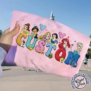 May include: Light pink sweatshirt with the word "CUSTOM" in colorful, stylized letters, each letter featuring a Disney princess. The sweatshirt has a round, embroidered logo that says "Gildan Sweatshirt Light Pink".