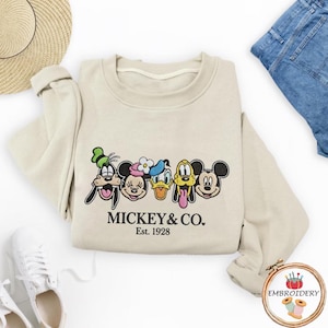 以下が含まれることがあります： ミッキーマウスと仲間たちの刺繍デザインが施されたベージュのスウェットシャツ。デザインにはグーフィー、ミニーマウス、ドナルドダック、プルート、ミッキーマウスが含まれています。「MICKEY & CO. Est. 1928」の文字が下に刺繍されています。