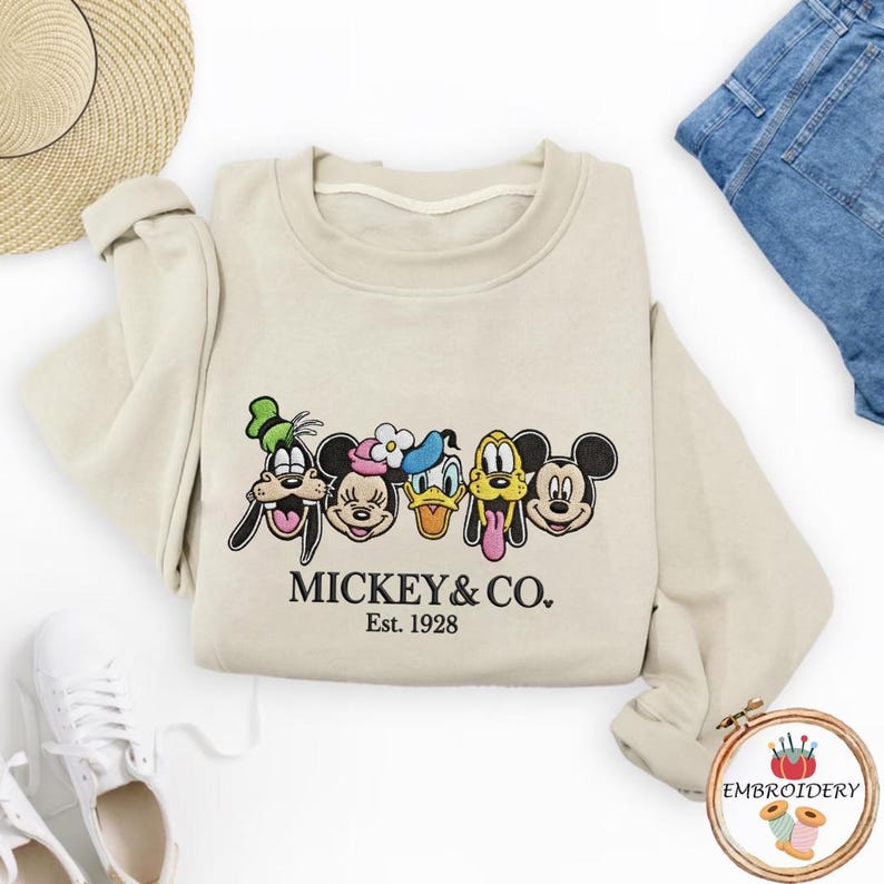 以下が含まれることがあります： ミッキーマウスと仲間たちの刺繍デザインが施されたベージュのスウェットシャツ。デザインにはグーフィー、ミニーマウス、ドナルドダック、プルート、ミッキーマウスが含まれています。「MICKEY & CO. Est. 1928」の文字が下に刺繍されています。