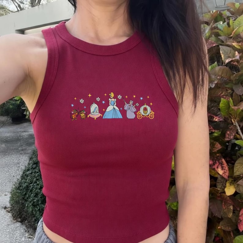 May include: A burgundy ribbed tank top with a crew neck. The front features an embroidered design of Cinderella and her iconic accessories, including mice, a glass slipper, and a carriage. The design is in shades of blue, yellow, and purple.