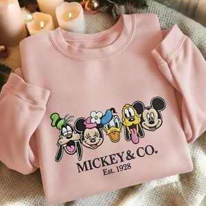 以下が含まれることがあります： ミッキーマウス、ミニーマウス、ドナルドダック、グーフィー、プルートの刺繍が施された薄いピンク色のスウェットシャツ。「MICKEY & CO. Est. 1928」の文字が下にプリントされています。スウェットシャツはクルーネックと長袖です。