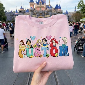 May include: A light pink sweatshirt featuring the word "CUSTOM" in colorful, cartoon-style letters, each letter adorned with a Disney princess. The princesses include Belle, Jasmine, Rapunzel, Snow White, Ariel, and Elsa. The background shows a theme park.