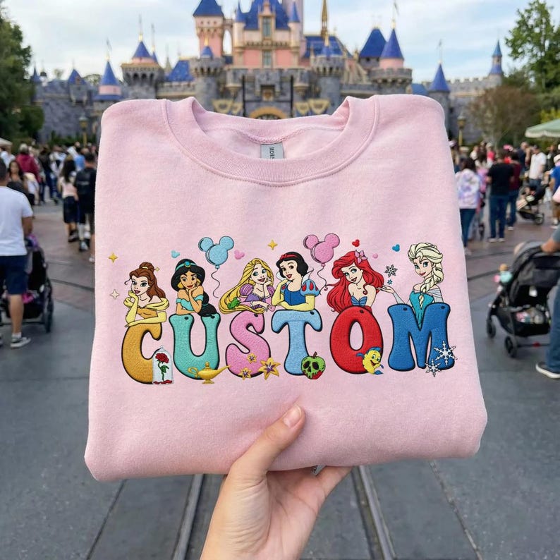 May include: A light pink sweatshirt featuring the word "CUSTOM" in colorful, cartoon-style letters, each letter adorned with a Disney princess. The princesses include Belle, Jasmine, Rapunzel, Snow White, Ariel, and Elsa. The background shows a theme park.