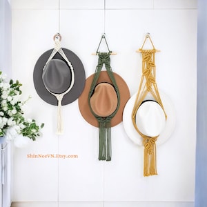 May include: Three macrame hat hangers suspended from hooks, featuring a cream hanger with a grey hat, an olive green hanger with a tan hat, and a mustard yellow hanger with a white hat. Each hanger includes knotted cord and a tassel.