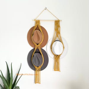 May include: A mustard yellow macrame wall hanging featuring two vertical sections suspended from a wooden dowel. The left section holds a tan hat and a dark grey hat, while the right section holds a white hat. The text ShinNeeVN.Etsy.com is visible.