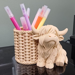 Op de afbeelding: Een beige pennenhouder met een gevlochten mandontwerp, naast een beige Highland koeienbeeldje. De pennenhouder bevat verschillende kleurrijke stiften. De items staan op een reflecterend zwart oppervlak.