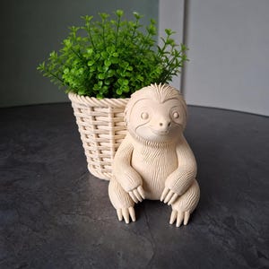 Op de afbeelding: Een beige keramieken luiaardbeeldje staat voor een kleine gevlochten mand met groene bladeren. De luiaard heeft een lachend gezicht en een gedetailleerde vachttextuur. De plantenbak en de luiaard staan op een donkergrijze ondergrond.