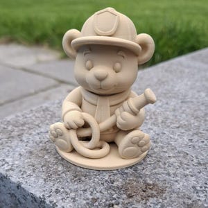 Peut inclure: Figurine d'ours beige portant un casque et une écharpe de pompier, tenant un tuyau d'arrosage. L'ours est assis, les jambes croisées. La figurine est faite d'un matériau solide et est posée sur une surface en pierre grise.