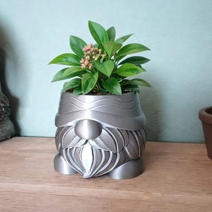 May include: A silver gnome-shaped planter holding a small green plant with pink flowers. The planter has a detailed beard and hat design. The plant pot is sitting on a wooden surface.