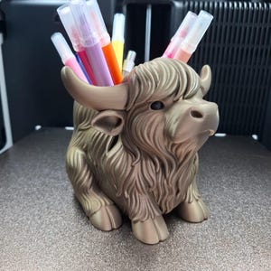 Op de afbeelding: Een Highland-koe-vormige pennenhouder in een bruine kleur, gevuld met kleurrijke stiften. De pennenhouder heeft een gedetailleerd, gestructureerd ontwerp, inclusief hoorns en harige vacht. De stiften zijn in verschillende kleuren, waaronder roze, paars, oranje en geel.