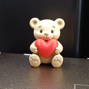 Op de afbeelding: Een crèmekleurig teddybeerbeeldje met een helderrood hart. De beer heeft kleine zwarte ogen en een schattige, vriendelijke uitdrukking. Het beeldje staat op een getextureerd oppervlak, met een donkere achtergrond.