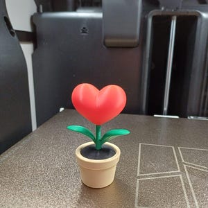 Op de afbeelding: Een 3D-geprinte decoratieve bloem met een rode hartvormige bloem, groene bladeren en een beige pot. De bloem staat tegen een neutrale achtergrond, waardoor de levendige kleuren en het unieke ontwerp van het handgemaakte item worden benadrukt.