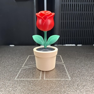 Op de afbeelding: Een 3D-geprinte rode roos in een beige pot met groene bladeren. De roos staat in volle bloei en de pot staat op een getextureerd oppervlak. De bloem is een decoratief item.