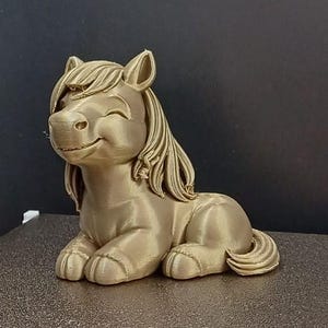 Könnte beinhalten: Eine goldfarbene, 3D-gedruckte Figur eines lächelnden Ponys in Ruheposition. Das Pony hat eine fließende Mähne und einen Schweif mit detaillierten Merkmalen. Die Figur ist wahrscheinlich ein Dekorationsartikel oder ein Spielzeug.