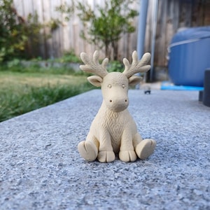 May include: A small, beige reindeer figurine with detailed antlers and a textured body. The reindeer is sitting on a gray surface, with a blurred background of greenery and a wooden fence.