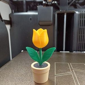 Peut inclure: Une tulipe jaune imprimée en 3D avec des feuilles et une tige vertes, dans un pot beige. La fleur est en pleine floraison et le pot a un intérieur gris foncé. L'objet est sur une surface texturée.