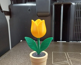 May include: A 3D-printed yellow tulip flower with green leaves and stem, set in a beige pot. The flower is in full bloom, and the pot has a dark gray interior. The object is on a textured surface.