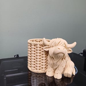Może przedstawiać: Beżowa figurka krowy rasy Highland obok pojemnika w stylu koszyka. Krowa ma szczeg&oacute;łowe futro i zakrzywione rogi. Kosz ma teksturowany, tkany wz&oacute;r. Przedmioty są prawdopodobnie dekoracyjne lub funkcjonalne.
