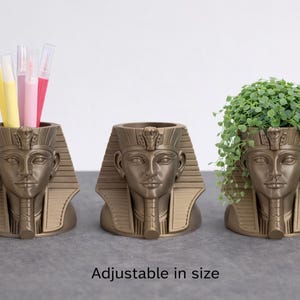 Op de afbeelding: Drie goudkleurige, Egyptische farao-hoofd-vormige plantenbakken. Eén bevat pennen, een andere is leeg en de derde is gevuld met groen. De tekst "Adjustable in size" staat onderaan.