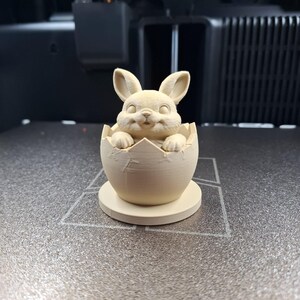 Op de afbeelding: Een 3D-geprint paashaasbeeldje dat uit een gebarsten eierschaal komt. Het konijn heeft lange oren en een lachend gezicht. Het ei en de basis zijn lichtbeige, geplaatst op een grijs oppervlak.