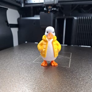 Puede incluir: Figura de pato impresa en 3D con cuerpo blanco, patas y pico naranjas y una chaqueta acolchada amarilla. El pato está sobre una superficie gris texturizada, con una impresora 3D en el fondo.