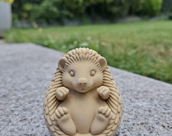 Puede incluir: Figura de erizo de color beige con detalles de púas, patas y una cara sonriente. La figura está sobre una superficie gris, con un fondo verde borroso de hierba y follaje.