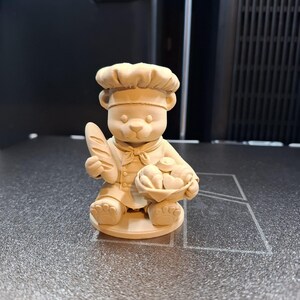 Op de afbeelding: Lichtbruin berenbeeldje als bakker, met een stokbrood en een mandje met gebakken goederen. De beer draagt een koksmuts en schort. Het beeldje staat op een getextureerd oppervlak.
