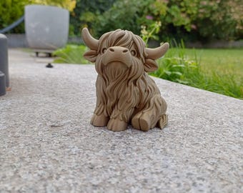 Może przedstawiać: Szczegółowa, brązowa figurka krowy rasy Highland, z długim, szorstkim futrem i zakrzywionymi rogami. Rzeźba stoi na betonowej powierzchni, z rozmytym zielonym tłem z liści i ogrodu.