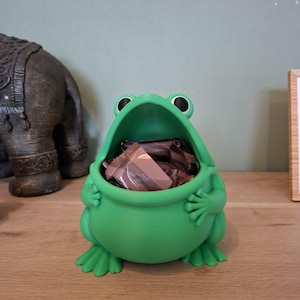 Op de afbeelding: Een felgroene kikker-vormige container gevuld met verpakte chocolade snoepjes. De container heeft grote, ronde ogen en een wijd open mond. De kikker staat op een houten oppervlak.