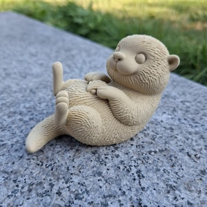 Op de afbeelding: Beige beeldje van een otter die op zijn rug ligt, poten op zijn borst. De otter heeft een textuur en ligt op een grijs gespikkelde ondergrond. Het beeldje is buiten.
