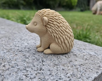 Puede incluir: Figura de erizo de color beige con púas detalladas, orejas pequeñas y pies diminutos. El erizo está sentado sobre una superficie de piedra gris, con un fondo verde borroso de hierba y follaje.