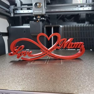 Op de afbeelding: Een rood 3D-geprint decoratief stuk met de woorden "Love Mom" verweven met een hart en een oneindigheidssymbool. Het stuk wordt weergegeven op een 3D-printerbed. De tekst is in cursief.