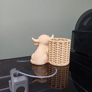 Może przedstawiać: Beżowa ceramiczna figurka krowy Highland i pleciony kosz są prezentowane na odblaskowej czarnej powierzchni. Szare urządzenie z napisem "Bambu Lab" jest r&oacute;wnież obecne, wraz z przezroczystą rurką.