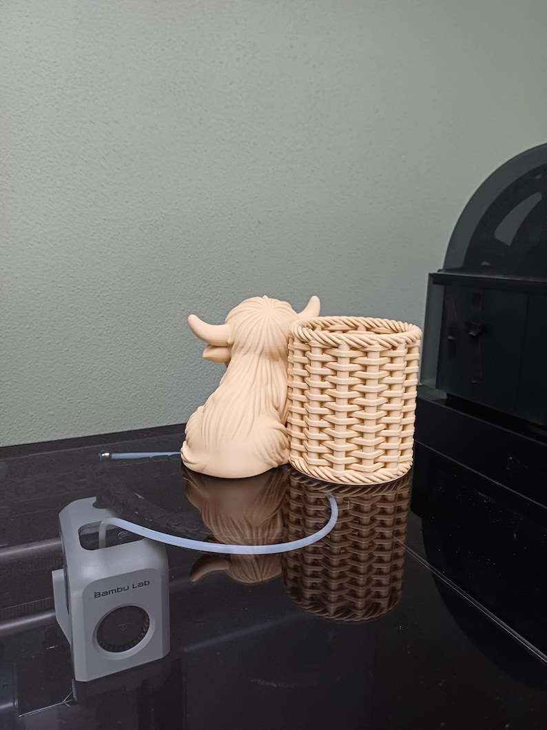 Może przedstawiać: Beżowa ceramiczna figurka krowy Highland i pleciony kosz są prezentowane na odblaskowej czarnej powierzchni. Szare urządzenie z napisem "Bambu Lab" jest r&oacute;wnież obecne, wraz z przezroczystą rurką.