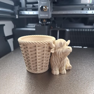 Könnte beinhalten: Ein hellbeige 3D-gedruckter Pflanztopf mit einem geflochtenen Korbmuster, neben einer Figur einer Highland-Kuh. Die Kuh ist ebenfalls hellbeige und hat langes, zotteliges Fell. Der Topf und die Kuh stehen auf einer grauen Oberfläche.