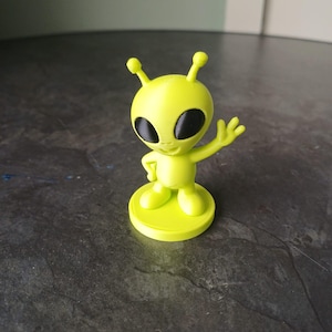 May include: A small, lime green alien figurine with large black eyes and a welcoming wave. The alien has two antennae and is standing on a circular base. The figurine is made of a smooth, solid material.