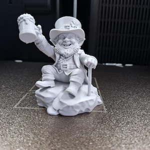 Puede incluir: Figura gris claro de un duende, sentado en una roca, sosteniendo una jarra de cerveza y un bastón. El duende lleva un sombrero con un trébol, una chaqueta y un cinturón. La figura es una impresión 3D.