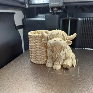 Może przedstawiać: Beżowa figurka krowy rasy Highland wydrukowana w 3D z pasującym, plecionym koszem. Krowa ma szczeg&oacute;łowe futro i rogi. Kosz jest cylindryczny z plecionym wzorem. Obiekty znajdują się na teksturowanej powierzchni, prawdopodobnie w drukarce 3D.