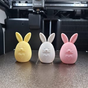 Op de afbeelding: Drie paashaasbeeldjes in geel, wit en roze. Elke konijn heeft lange oren, een hart en snorharen. De konijnen staan op een grijs oppervlak, mogelijk een 3D-printerbed.